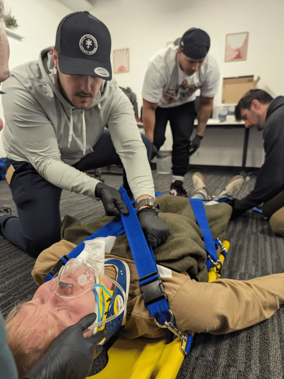 Four students packaging a patient on a spine board: collar and head blocks in place, oxygen mask on, straps being tightened by a student in a Delta cap